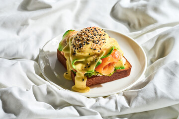 Toast with a poached egg, hollandaise sauce and salmon and avocado in a plate on a white sheet. Tough light.