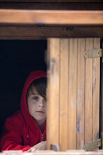 Preteen Boy In Red Sweatshirt, Hiding Behind A Wooden Door, Looking Scared
