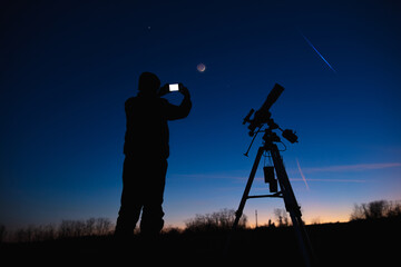 silhouette of a man with smartphone, telescope and countryside under evening skies.