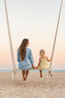 © somemeans - Young mother and little fair-haired girl sit on swing on sea background during sunset. Mom and daughter on the beach over yellow magic sunset lights.