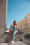 Fototapeta Miasto - young curly brunette girl in white top and blue jeans is sitting casual near street store background at sunny day and looking away with smile. lifestyle concept, free space