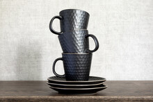 Three Dark Ceramic Tea Or Coffee Mugs Standing On Top Of Each Other. Dark Modern Cups Stacked On A Wooden Background