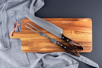 Fork and knife for meat on the wooden board with grey apron on black background