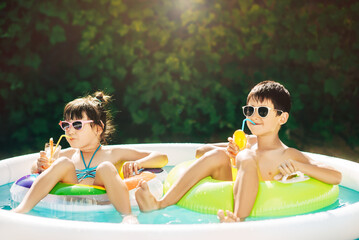 Two young children, boy and girl, sibling, resting in summer in inflatable pool. Children swim in inflatable circles and drink orange juice from tubes. Sunny summer day. Happy summer vacation.