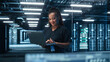 © Gorodenkoff - Portrait of Successful African American Female IT Specialist Using Laptop, Standing in Data Center. System Administrator Works on Web Services. Cloud Computing, Server Analytics, Cyber Security