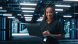 © Gorodenkoff - African American Female IT Specialist Using Laptop Computer, Standing in Big Warehouse Data Center. System Administrator working with SAAS, Cloud Web Services. E-Business Digital Entrepreneur