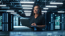 Portrait Of Multiracial Mature Female IT Specialist Using Laptop, Standing In Data Center. Infrastructure Architect Works On Web Services. Cloud Computing, Server Analytics, Cyber Security Maintenance