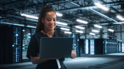 Poster - Multiracial Data Center IT Technician Standing at the Server Rack Corridor with a Laptop Computer. She is Visually Inspecting Something while Looking Away. Night Office Concept
