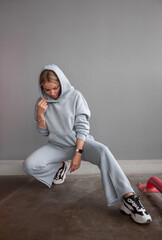 Wall Mural - blonde girl in gray oversize hoodie and gray pants sits fashion near gray wall background at parking and looks down with hand near hood. lifestyle concept, free space