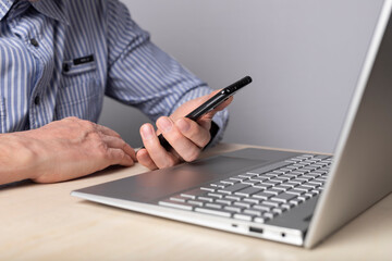Poster - Man sitting at table with laptop and holding phone. Checking information, reading or typing message while working on computer. Man hands closeup. High quality photo