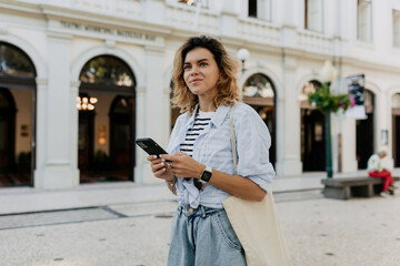 Outside portrait of pretty woman with wavy hair wearing striped t-shirt and blue shirt with eco bag walking on the street in sunny warm day and using smartphone