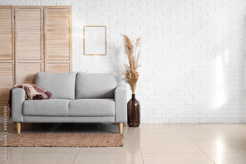 Stylish sofa and vase with dry reeds near white brick wall