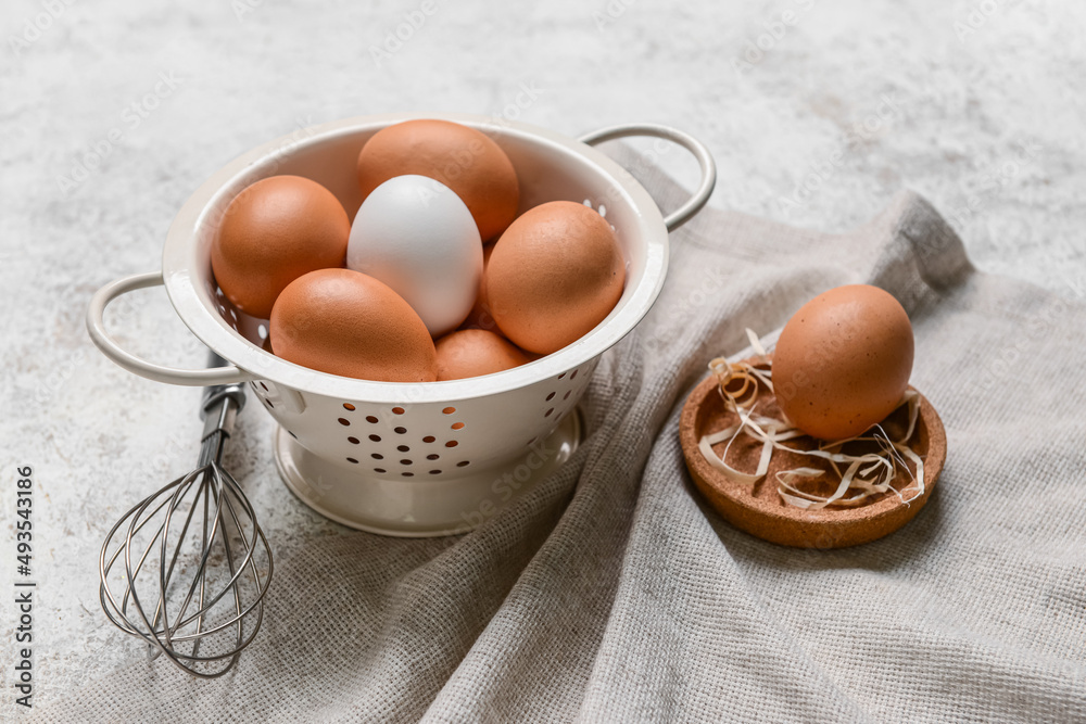 Colander with fresh chicken eggs and whisk on light background