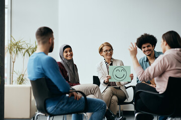 Wall Mural - Multiracial people having group therapy with mental health professional who is holding placard with drawn smiley face.