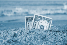 Two One Dollar Bills Half Buried In Sand On Sandy Seashore Close-up. Concept Money, Finance, Investment Wealth Poverty, Financing, Cash. Dollar Bills Half In Sand On Sea Beach On Sunny Day. Blue Color