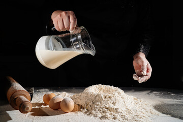 woman pours milk from a jug into flour to knead the dough on black background. 