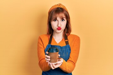 Wall Mural - Redhead young woman wearing waitress apron holding take away cup of coffee puffing cheeks with funny face. mouth inflated with air, catching air.