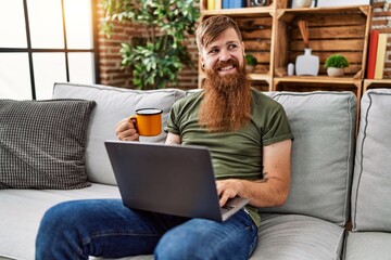 Wall Mural - Young redhead man drinking coffee and using laptop at home