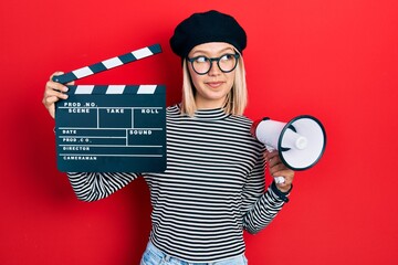 Poster - Beautiful blonde woman holding video film clapboard and megaphone smiling looking to the side and staring away thinking.