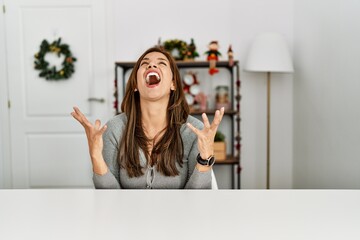 Poster - Young latin woman sitting on the table by christmas decor celebrating mad and crazy for success with arms raised and closed eyes screaming excited. winner concept