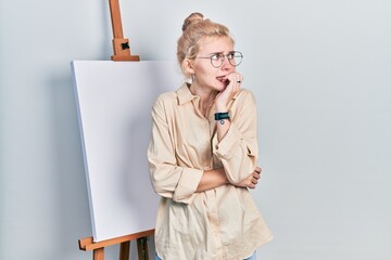 Poster - Beautiful caucasian woman with blond hair standing by painter easel stand looking stressed and nervous with hands on mouth biting nails. anxiety problem.