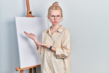 Poster - Beautiful caucasian woman with blond hair standing by painter easel stand presenting canva skeptic and nervous, frowning upset because of problem. negative person.