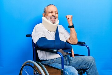 Canvas Print - Handsome senior man with beard sitting on wheelchair with neck collar an arm sling screaming proud, celebrating victory and success very excited with raised arm