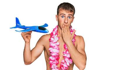 Wall Mural - Young blond man wearing swimsuit and hawaiian lei holding airplane toy covering mouth with hand, shocked and afraid for mistake. surprised expression