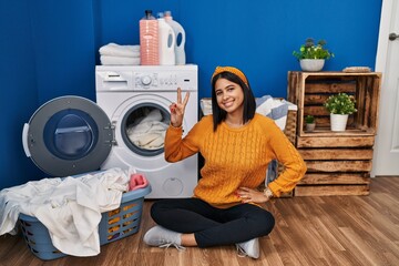 Poster - Young hispanic woman doing laundry smiling looking to the camera showing fingers doing victory sign. number two.