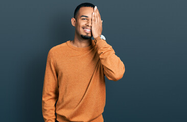 Wall Mural - Young african american man wearing casual clothes covering one eye with hand, confident smile on face and surprise emotion.