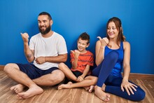 Family Of Three Sitting On The Floor At Home Smiling With Happy Face Looking And Pointing To The Side With Thumb Up.