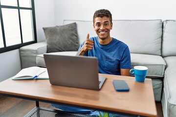 Wall Mural - Young handsome hispanic man using laptop sitting on the floor doing happy thumbs up gesture with hand. approving expression looking at the camera showing success.