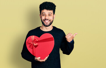 Poster - Young arab man with beard holding valentine gift pointing thumb up to the side smiling happy with open mouth