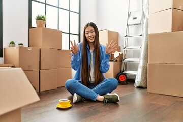 Canvas Print - Young chinese girl sitting on the floor at new home showing and pointing up with fingers number eight while smiling confident and happy.