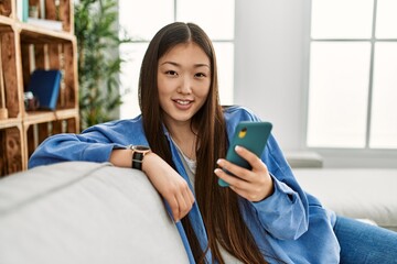 Wall Mural - Young chinese girl using smartphone sitting on the sofa at home.