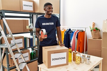 Poster - Young african american volunteer man packing donations box for charity looking away to side with smile on face, natural expression. laughing confident.