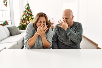 Sticker - Middle age hispanic couple sitting on the table by christmas tree smelling something stinky and disgusting, intolerable smell, holding breath with fingers on nose. bad smell