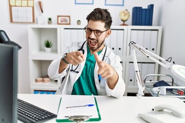 Poster - Young man with beard wearing doctor uniform and stethoscope at the clinic pointing fingers to camera with happy and funny face. good energy and vibes.