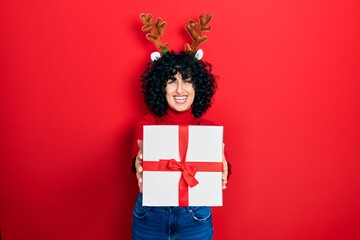 Sticker - Young middle east woman wearing deer christmas hat holding gift smiling and laughing hard out loud because funny crazy joke.