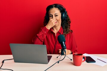 Poster - Beautiful middle age woman working at radio studio smelling something stinky and disgusting, intolerable smell, holding breath with fingers on nose. bad smell