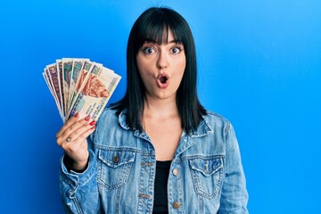 Wall Mural - Young hispanic woman holding egyptian pounds banknotes scared and amazed with open mouth for surprise, disbelief face