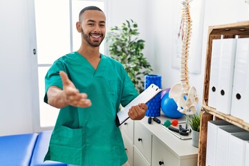 Wall Mural - African american physiotherapist man working at pain recovery clinic smiling cheerful offering palm hand giving assistance and acceptance.