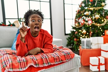 Canvas Print - Young african american woman lying on the sofa by christmas tree pointing finger up with successful idea. exited and happy. number one.