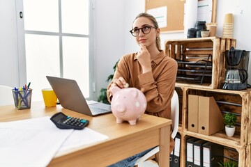 Canvas Print - Young blonde woman at the office putting coin into piggy bank serious face thinking about question with hand on chin, thoughtful about confusing idea