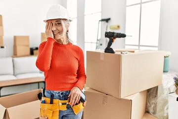 Poster - Middle age grey-haired woman wearing hardhat standing at new home covering one eye with hand, confident smile on face and surprise emotion.