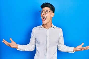 Sticker - Young hispanic man wearing casual clothes and glasses crazy and mad shouting and yelling with aggressive expression and arms raised. frustration concept.