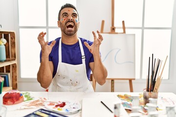 Sticker - Young hispanic man with beard at art studio with painted face crazy and mad shouting and yelling with aggressive expression and arms raised. frustration concept.