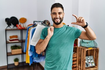 Sticker - Young handsome man with beard holding shopping bags at retail shop smiling and confident gesturing with hand doing small size sign with fingers looking and the camera. measure concept.