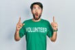 © Krakenimages.com - Young hispanic man wearing volunteer t shirt amazed and surprised looking up and pointing with fingers and raised arms.