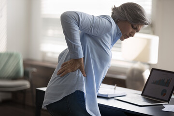 Rear side view aged woman stand near workplace desk, touch lower back, suffer from sudden backache, due to uncomfortable chair. Sedentary work disease, rheumatism, low-back lumbar pain illness concept